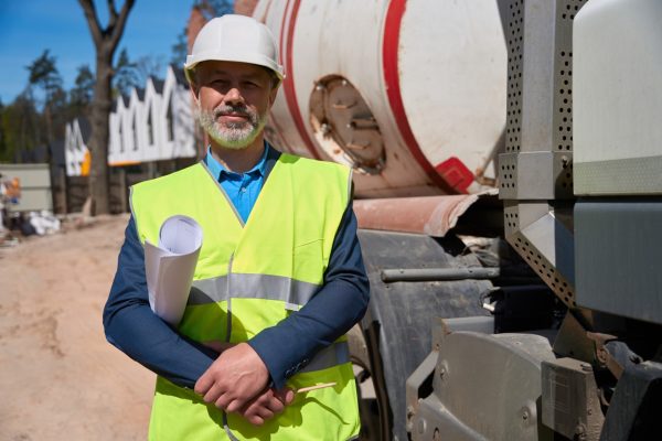 Main construction worker in hardhat and hi vis safety vest standing with blueprint in hands on background of concrete mixer truck